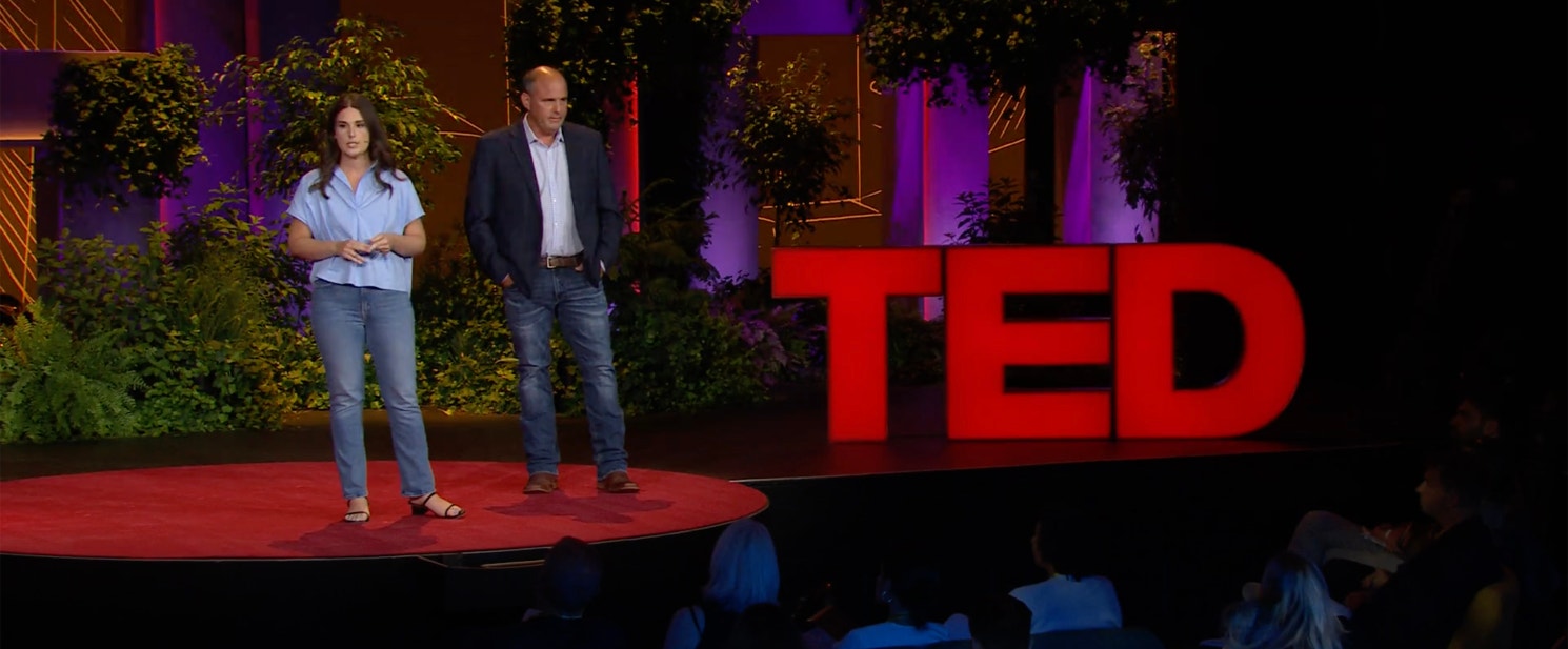 From left to right, Jessica and Jim Whitaker standing on a TED Talk stage next to the platform’s red logo.
